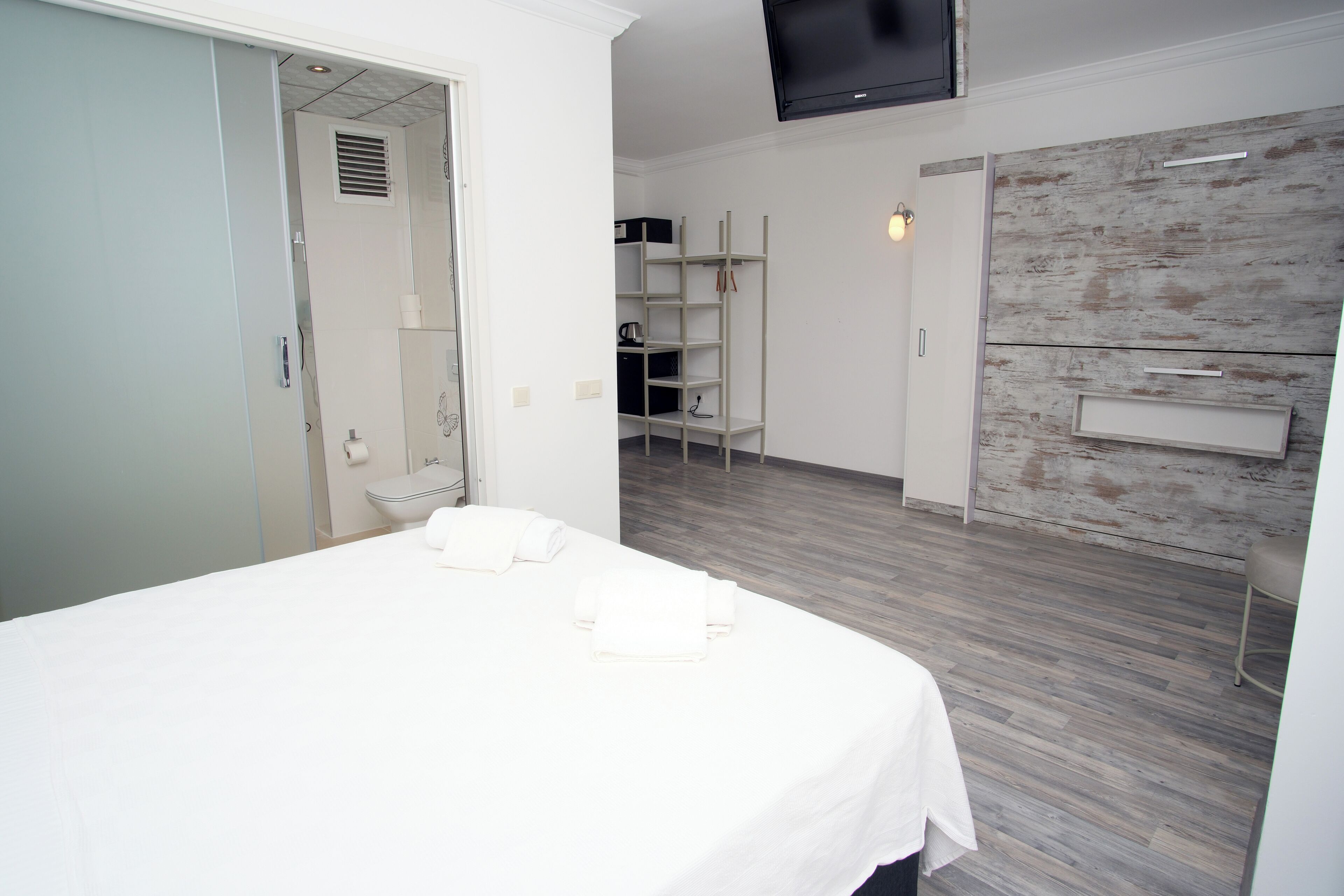 A modern hotel room with a large bed, a wooden floor, a built-in wardrobe, and a flat-screen TV mounted on the wall.