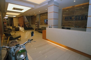 A hotel reception area with a reception desk, a staircase, and a seating area with chairs and a sofa.