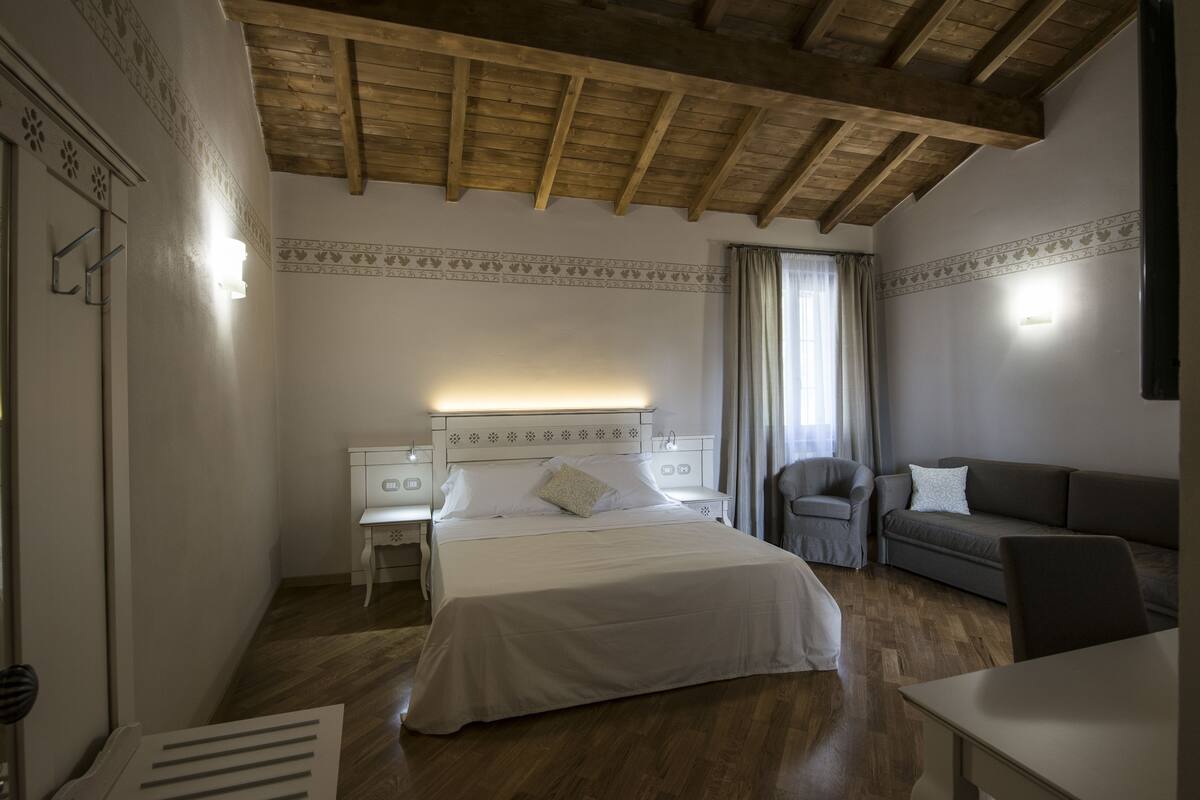 Un dormitorio con techo de madera, una cama con ropa blanca, un sofá gris y una pequeña mesa.