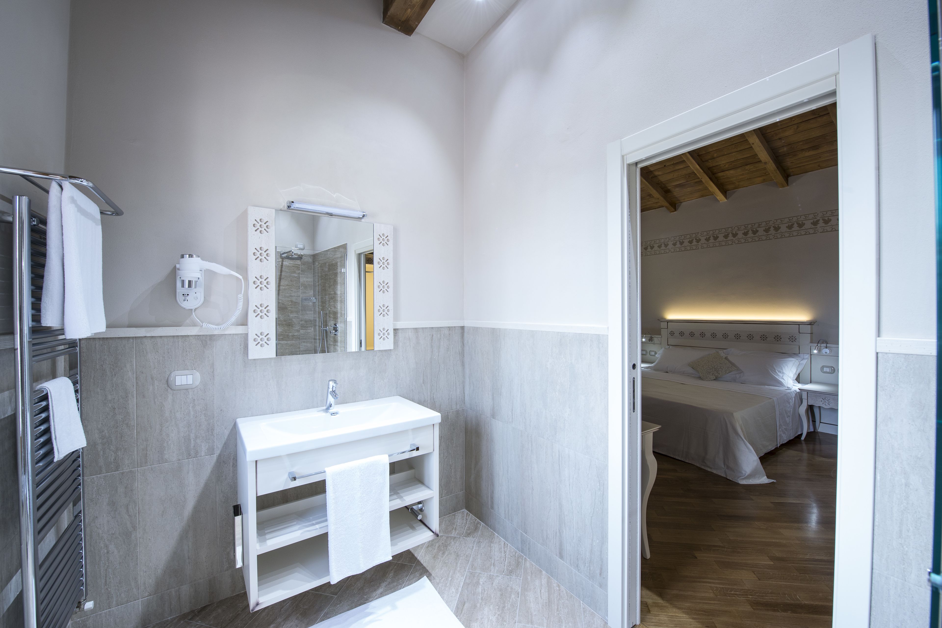 Ein modernes Badezimmer mit einem weißen Waschbecken, Spiegel und Handtuchhalter. Im Hintergrund ist eine Duschkabine zu sehen, und durch eine offene Tür gelangt man in einen Schlafbereich mit Bett und Nachttisch.