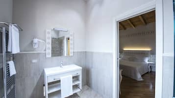 Ein modernes Badezimmer mit einem weißen Waschbecken, Spiegel und Handtuchhalter. Im Hintergrund ist eine Duschkabine zu sehen, und durch eine offene Tür gelangt man in einen Schlafbereich mit Bett und Nachttisch.