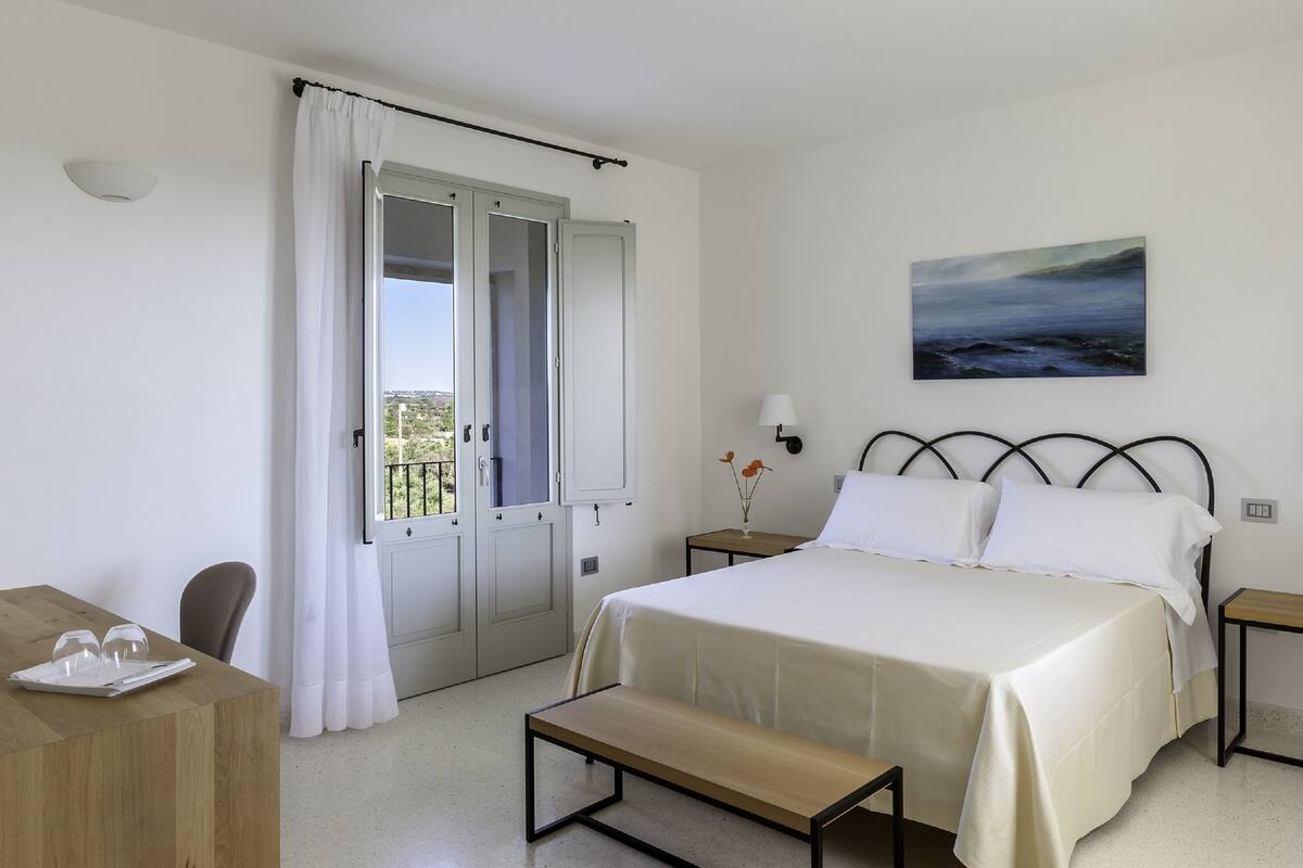 A bedroom with a bed, a bedside table, a bench, a wooden desk, and a window with curtains.