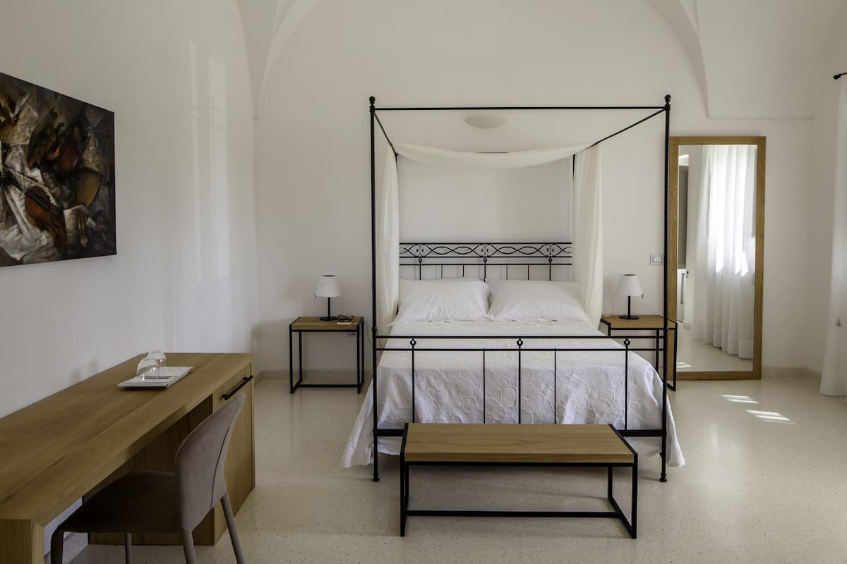 A four-poster bed with white linens, a wooden desk with a chair, a small bedside table with a lamp, and a large mirror on the wall.
