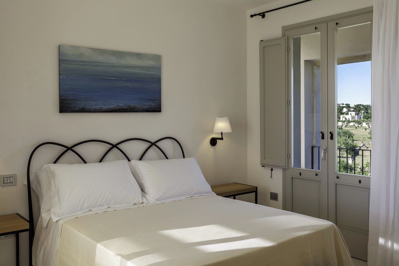 Un dormitorio con cama, mesita de noche, lámpara de pared, un cuadro y una ventana con puerta abierta.