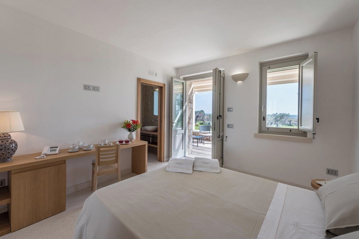 A hotel room with a bed, a desk with a phone and a vase of flowers, a bathroom visible through an open door, and a window with a view of the outdoors.
