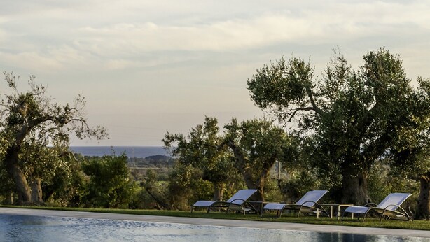 Un área junto a la piscina con un sofá blanco, una mesa con una botella y vasos, y sillas de tomar el sol.