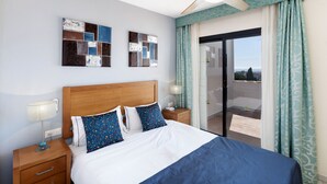 A hotel room with a bed, nightstands, a ceiling fan, artwork on the walls, and a view through the glass door. - Crown Resorts Club Calahonda (Mijas)