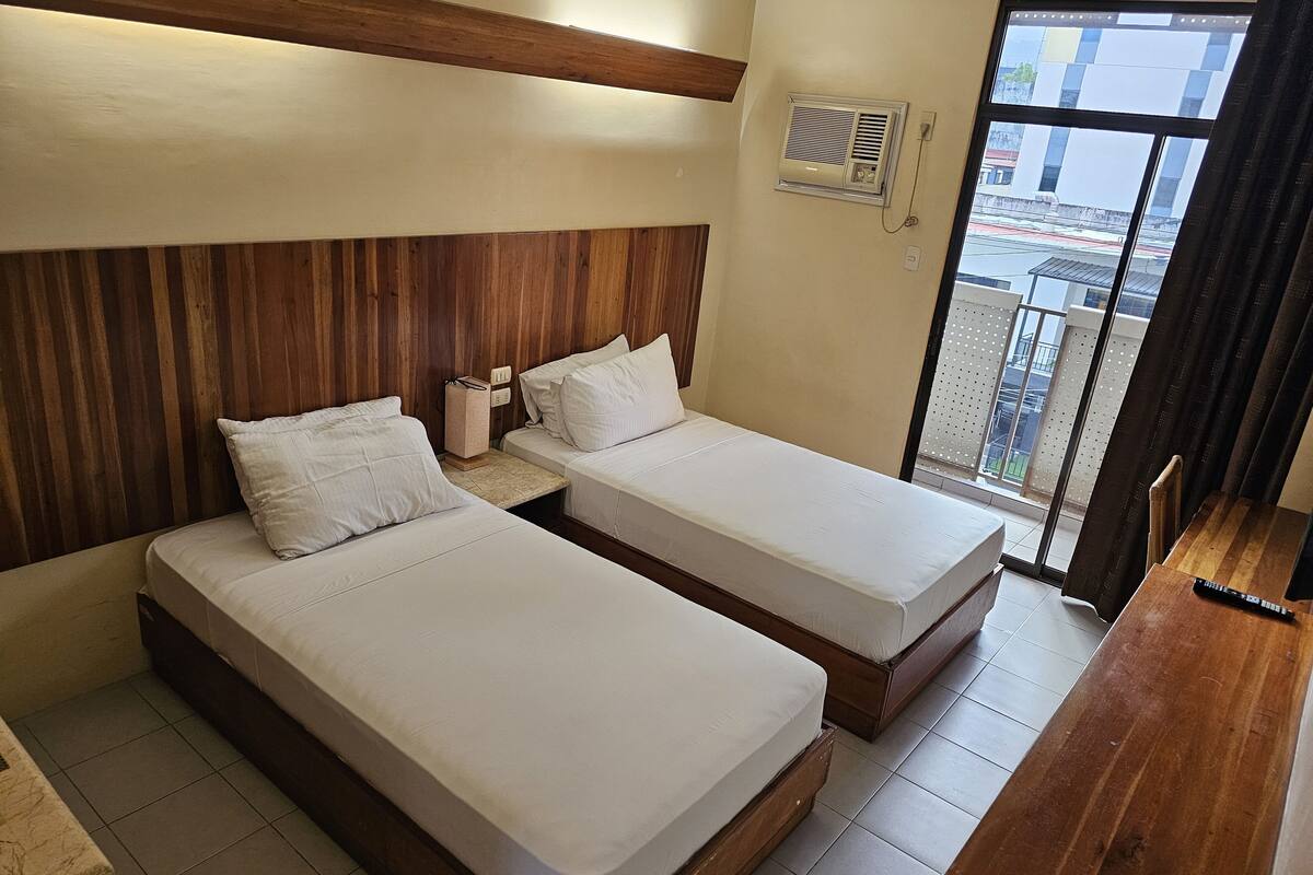 A hotel room with two single beds, a wooden headboard, a small bedside table with a lamp, and a window with a view of buildings.