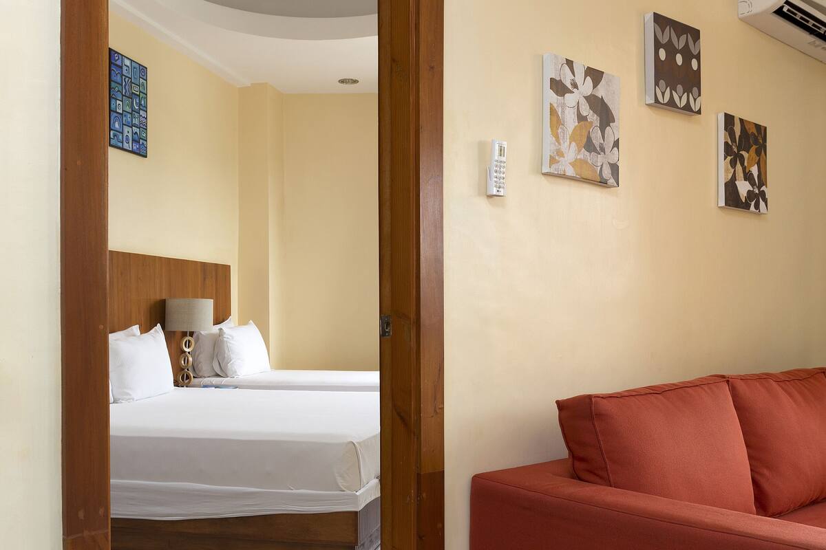 A hotel room with a wooden door leading to a bedroom with two beds and a red sofa.