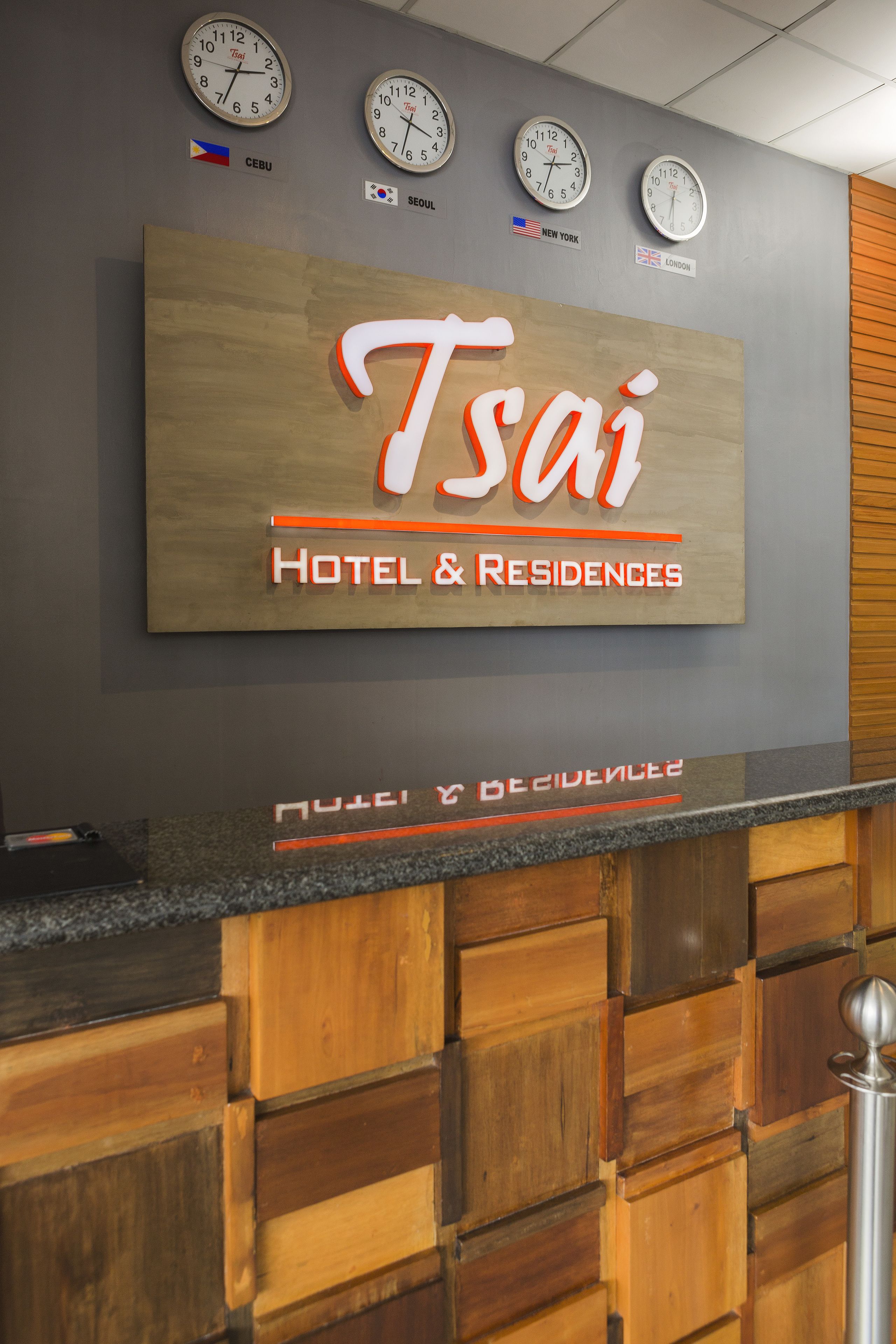 Un comptoir d’accueil avec une enseigne indiquant « Tsai Hotel & Residences », agrémenté d’horloges ornées de drapeaux de différentes villes.