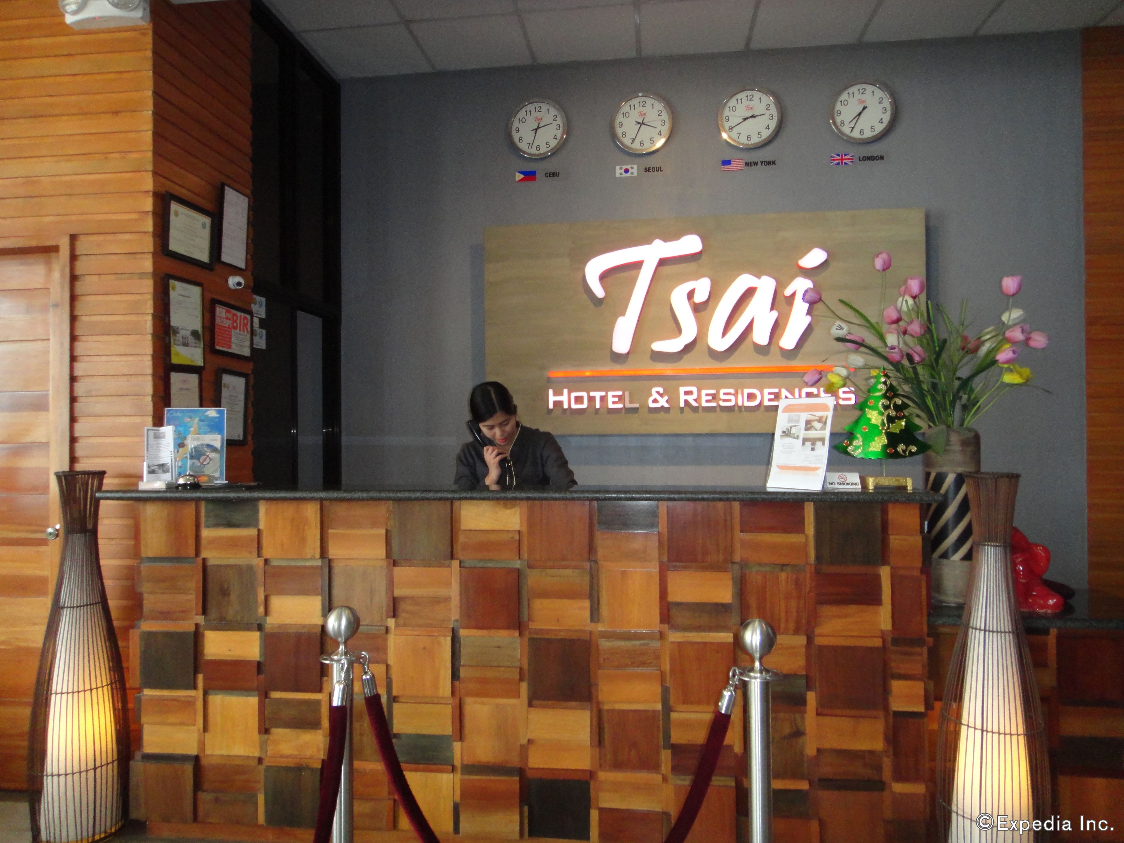 Tsai Hotel & Residences -hotellin vastaanottotiski, jossa on kelloja, jotka näyttävät eri aikavyöhykkeiden aikaa, vastaanottovirkailija ja koristeellinen maljakko, jossa on kukkia.