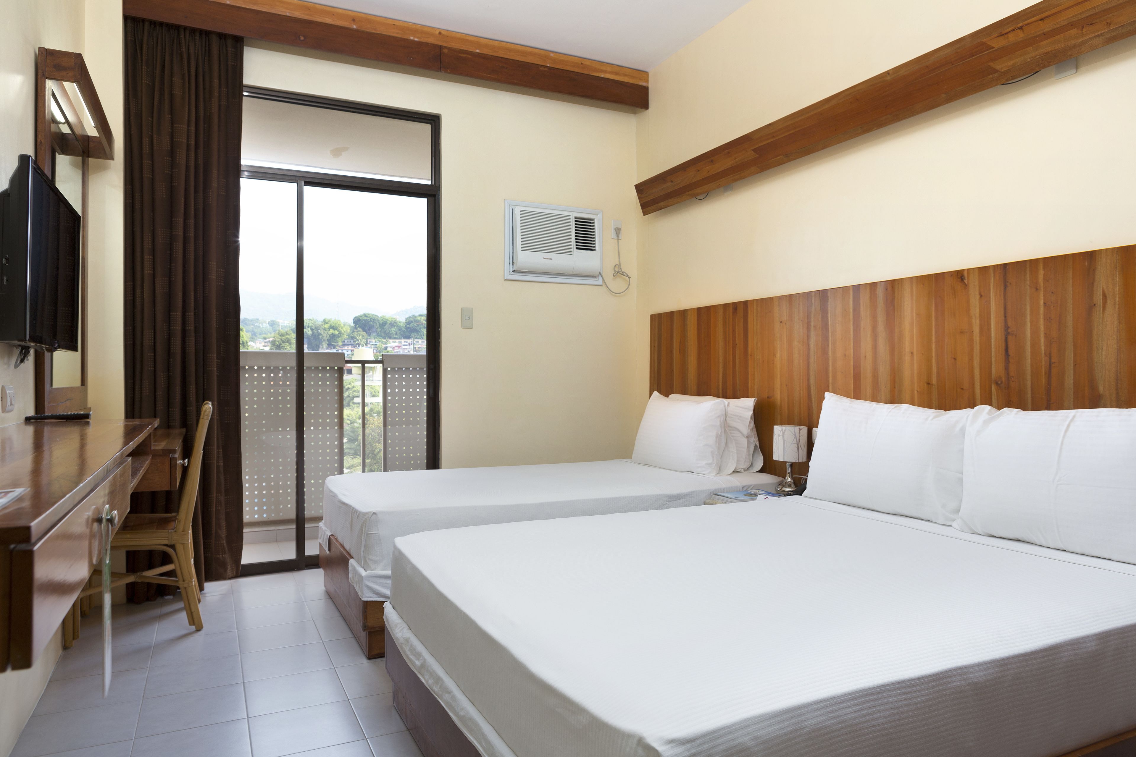 A hotel room with two beds, a desk, a chair, an air conditioning unit, and a view through a sliding door.