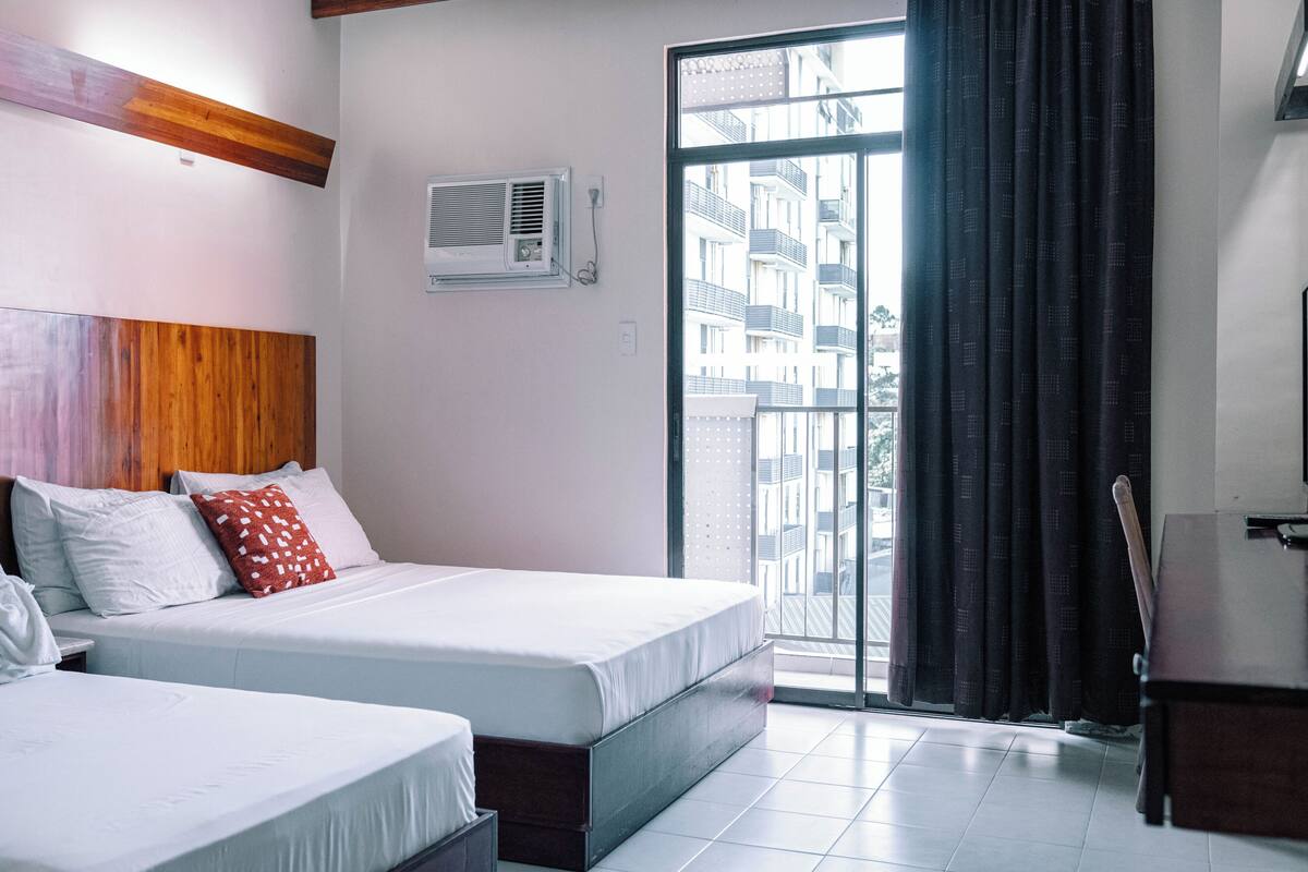 A hotel room with two beds, a wooden headboard, an air conditioning unit, a view of the city through a large window, and a dark curtain.