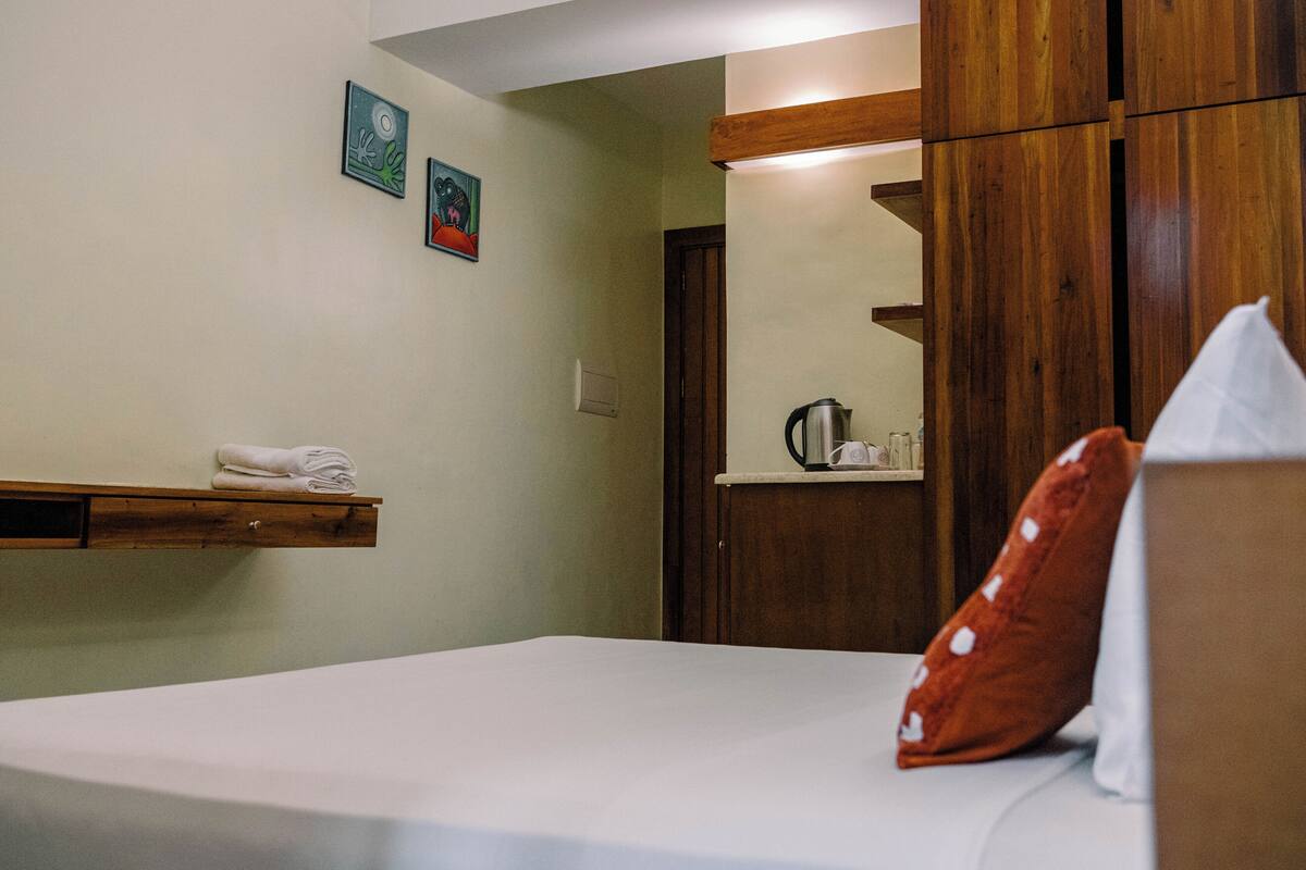 A bedroom with a bed, a wooden shelf with towels, a wooden wardrobe, and a small kitchenette area.