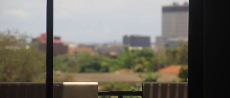 A view through a window with a metal railing, featuring a cityscape with buildings and greenery in the background.