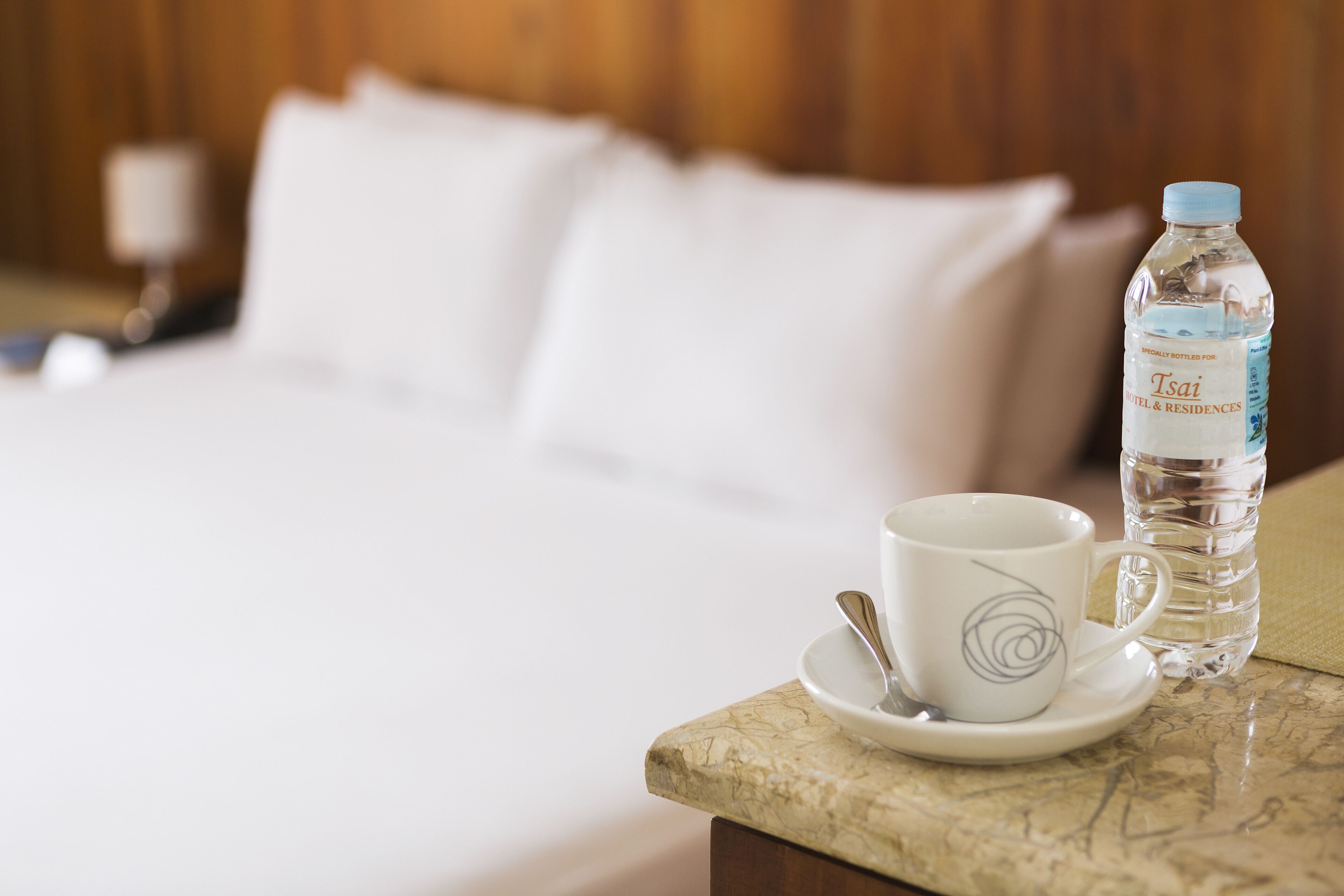 A neatly made bed with white linens, a water bottle, and a cup with a spoon on a marble table.