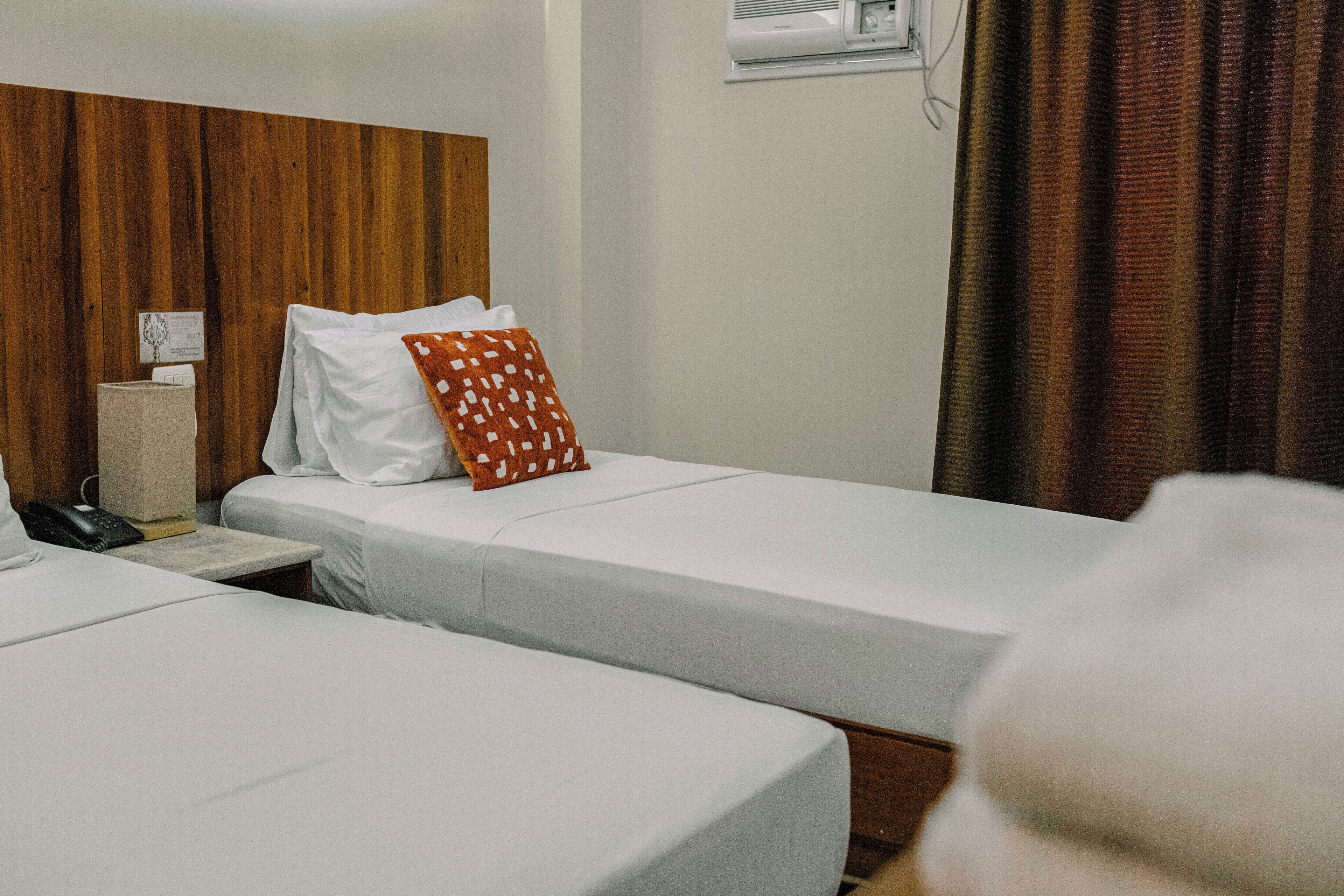 A hotel room with two single beds, a wooden headboard, a bedside table with a lamp, and a window with curtains.