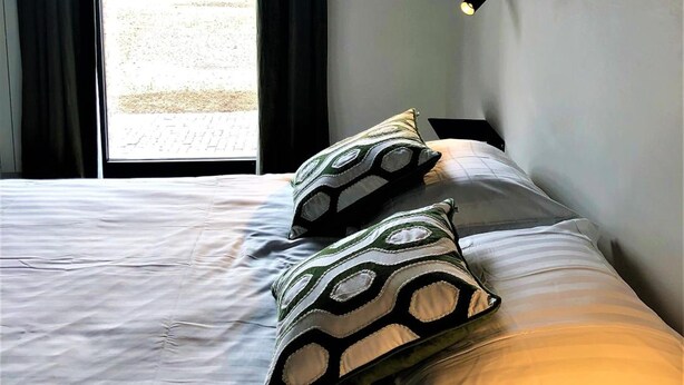 A hotel room with a bed, two patterned pillows, a window with curtains, and a wall-mounted light.