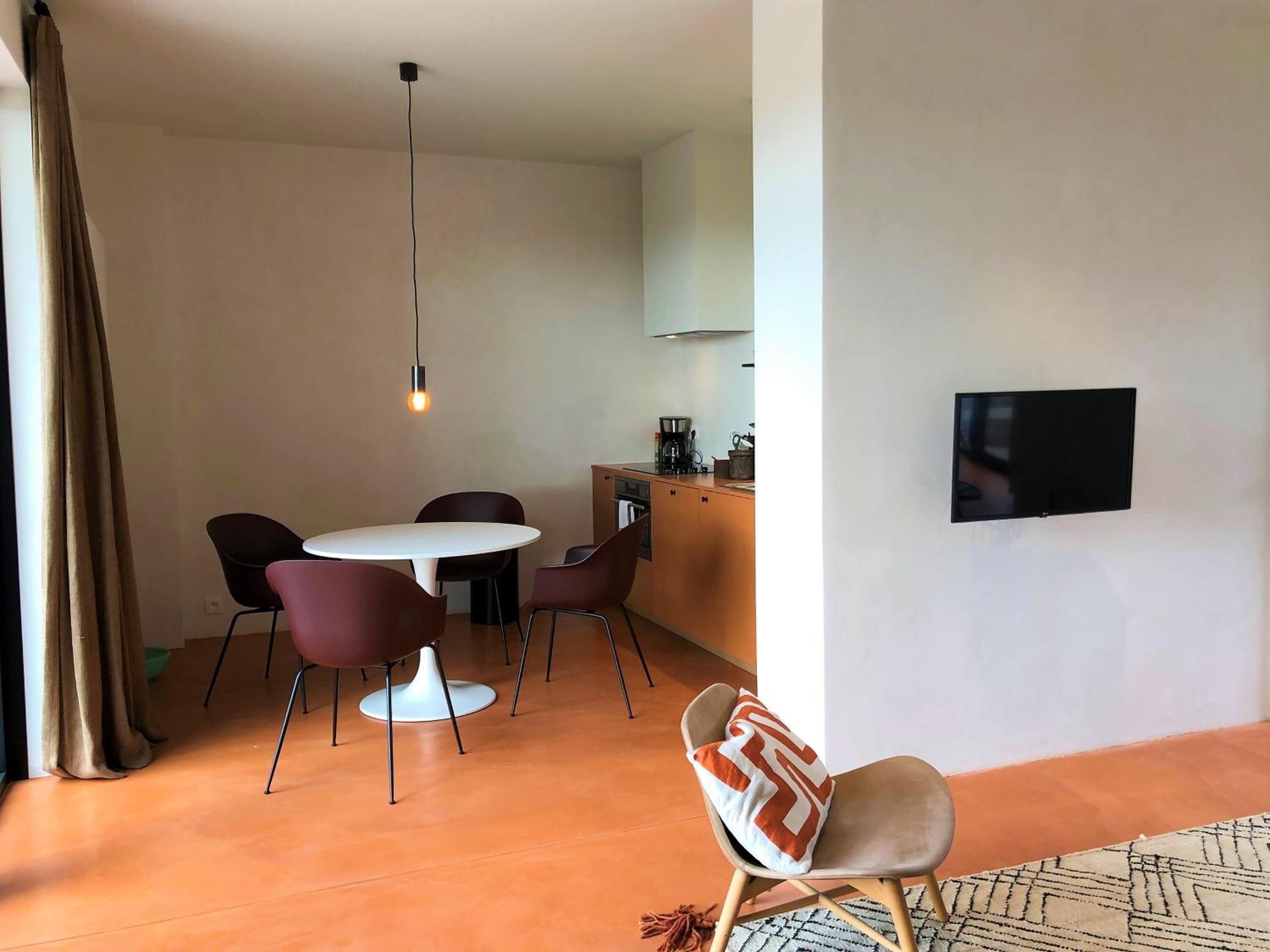 Een moderne woonkamer met een ronde eettafel, twee stoelen, een bank, een aan de muur bevestigde televisie en een keuken met een fornuis en kastjes.