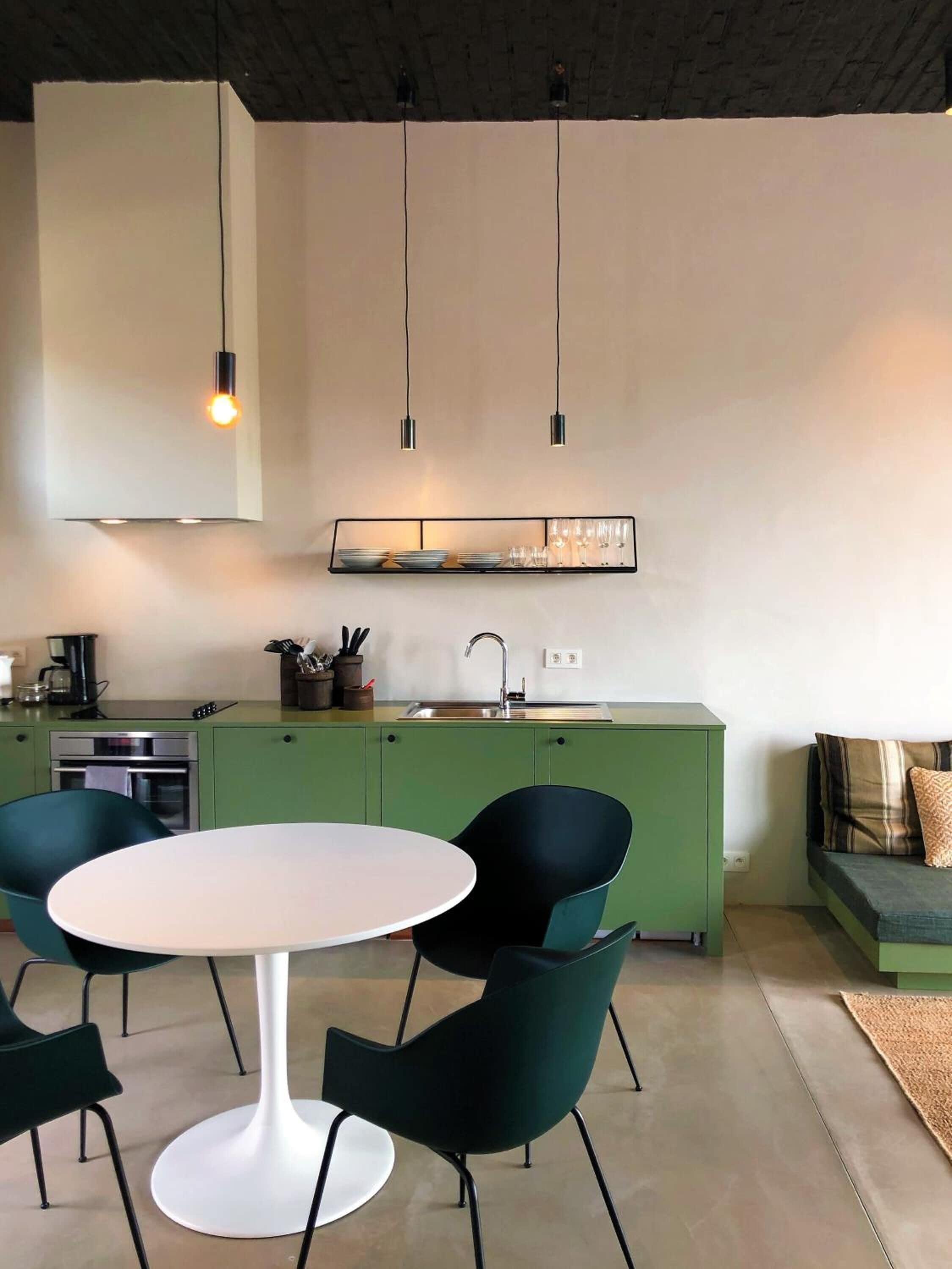 Een moderne keuken met groene kasten, een witte eettafel en zwarte stoelen. Boven de tafel hangen hanglampen en een andere hangende lamp.