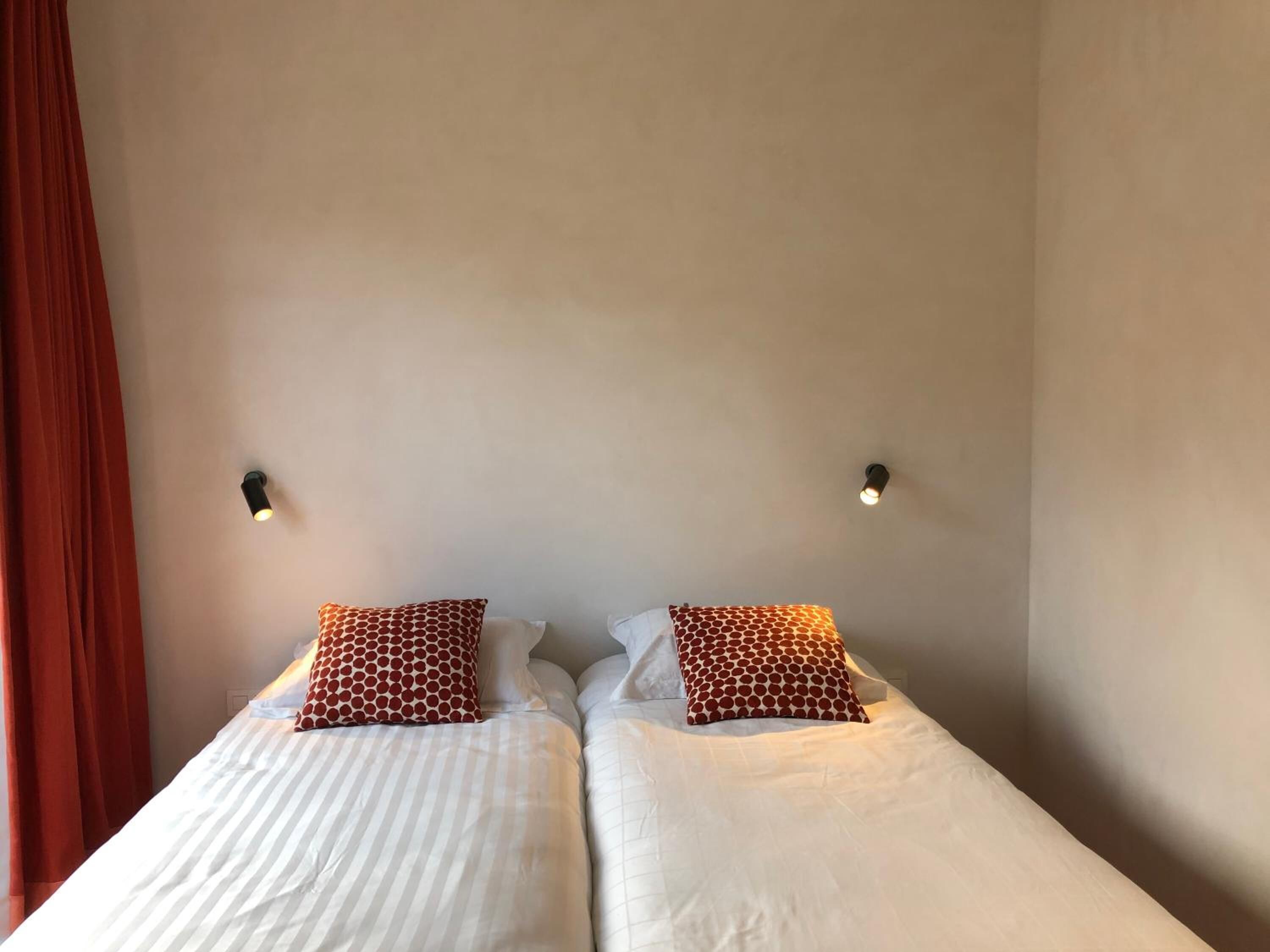 Cama doble con ropa de cama blanca y dos almohadones estampados, una cortina roja y dos luces fijadas a la pared.