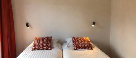 Cama doble con ropa de cama blanca y dos almohadones estampados, una cortina roja y dos luces fijadas a la pared.