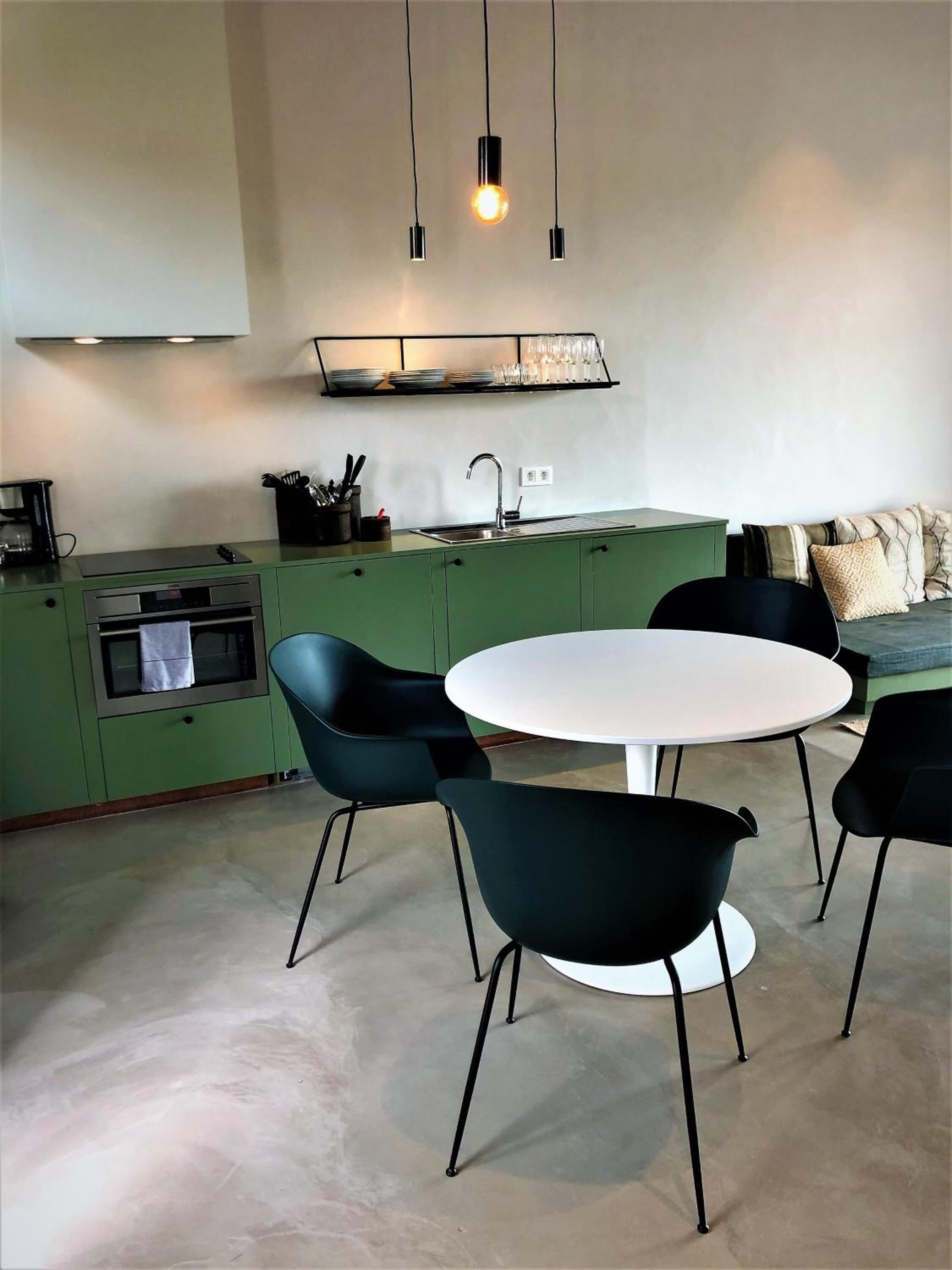 Una cocina moderna con armarios verdes, una mesa de comedor blanca y sillas negras. Se observa una cafetera, un lavamanos y una estantería con platos y vasos.