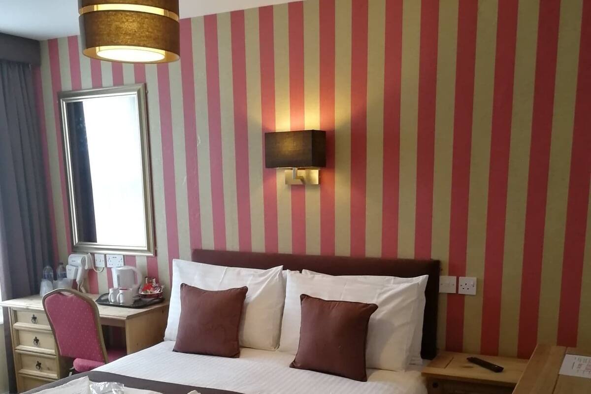 A hotel room with a large bed, a desk with a chair, a mirror, a wall-mounted lamp, and a striped wall.