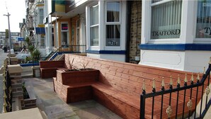 A wooden bench with a black metal railing, located on a sidewalk in front of a building with a sign that reads "TRAFFORD". - The Trafford Hotel (Blackpool)