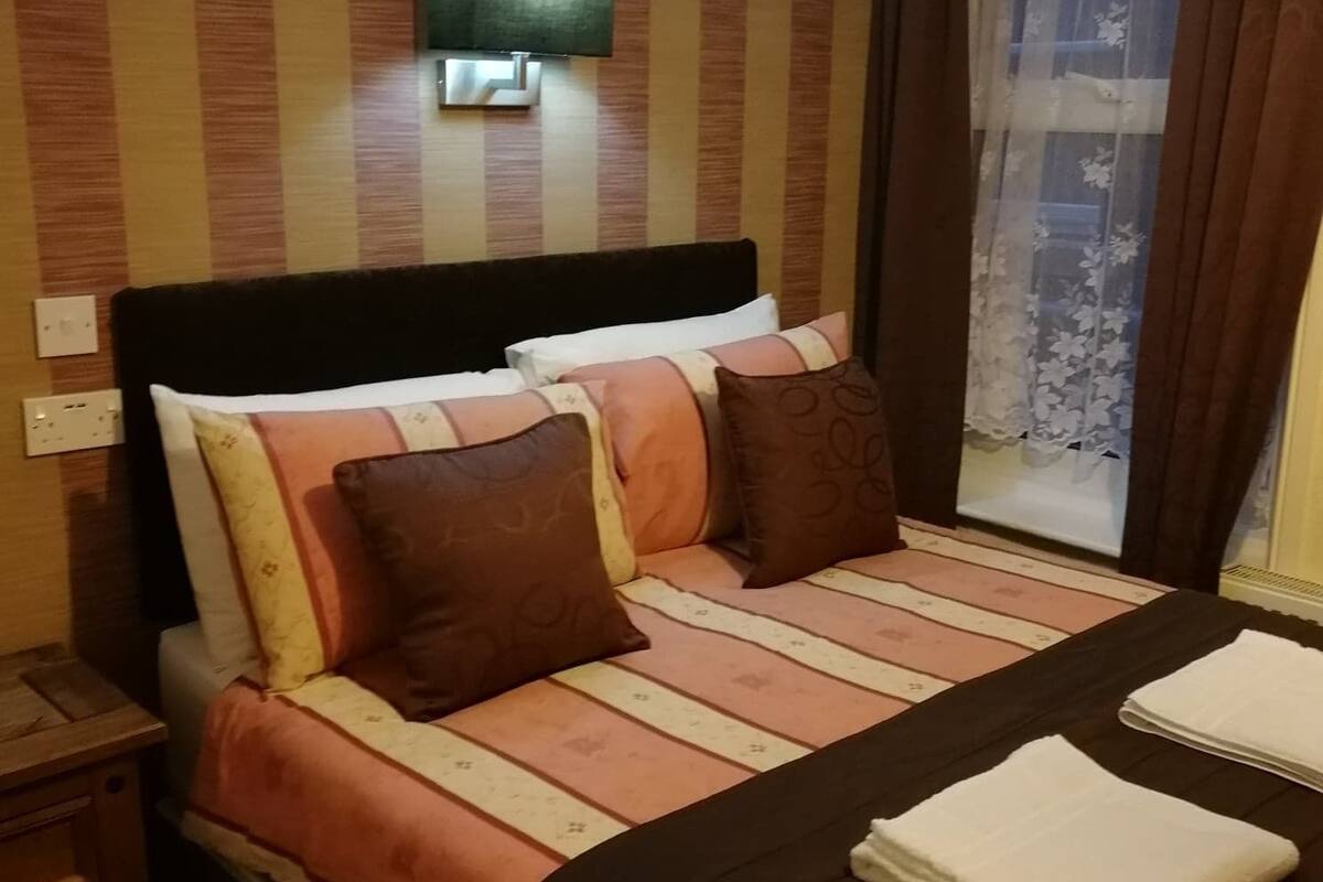 A hotel room with a bed, pillows, a nightstand, a lamp, and a window with curtains.