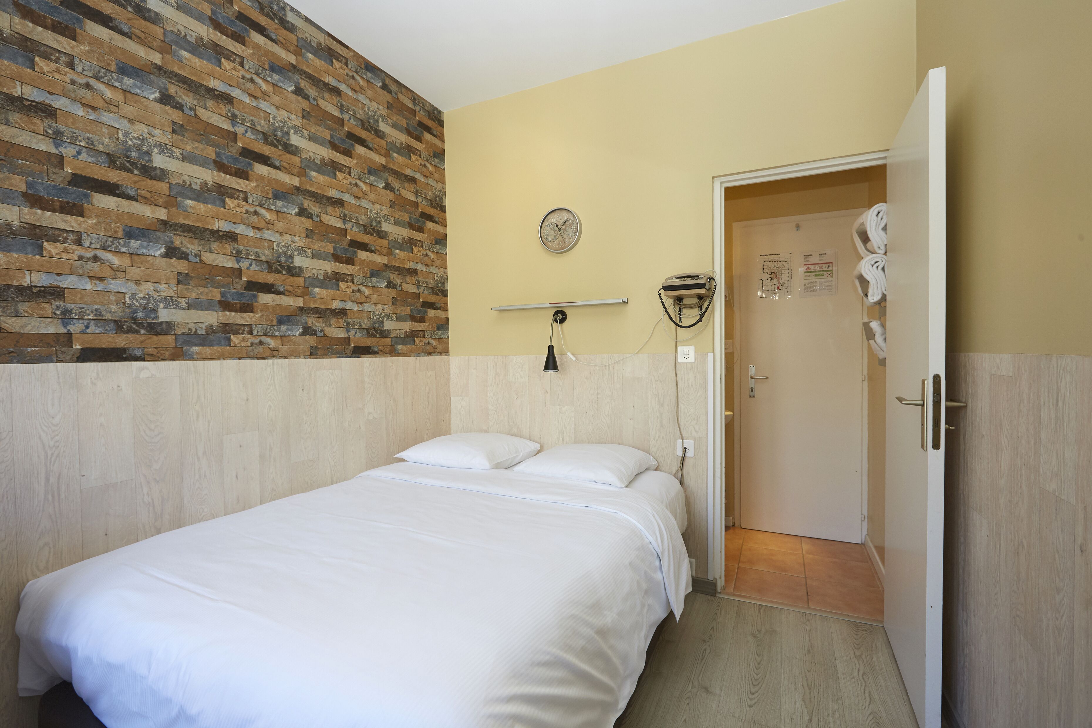 A hotel room with a bed, a brick accent wall, a clock, a towel rack, and a door leading to a bathroom.
