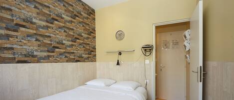 A hotel room with a bed, a brick accent wall, a clock, a towel rack, and a door leading to a bathroom.