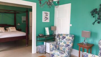 A room with a four-poster bed, a chair with a tropical print, a small table, a desk with a lamp, and a wall-mounted picture of birds.