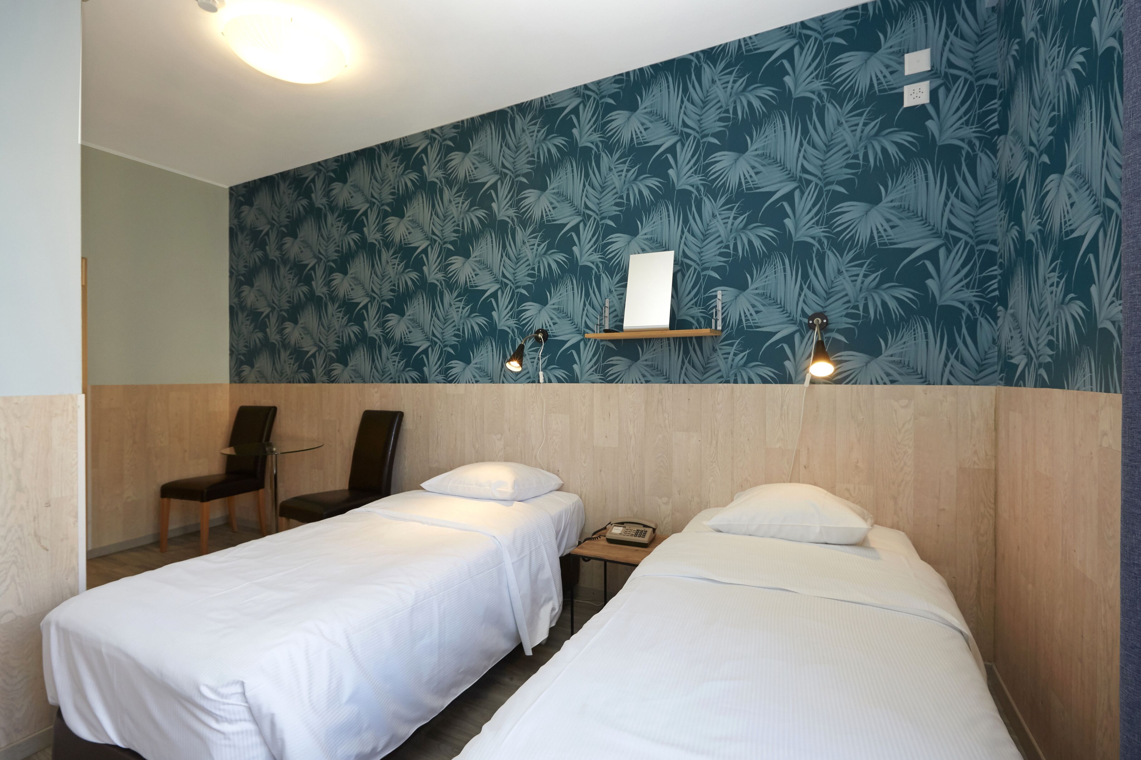 Une chambre d’hôtel avec deux lits simples, une tête de lit en bois, une petite table avec un téléphone, deux chaises, une étagère murale avec un tableau, et un luminaire au plafond.