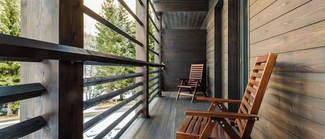 Una terraza de madera con barandilla, dos sillas de madera y vista a árboles.