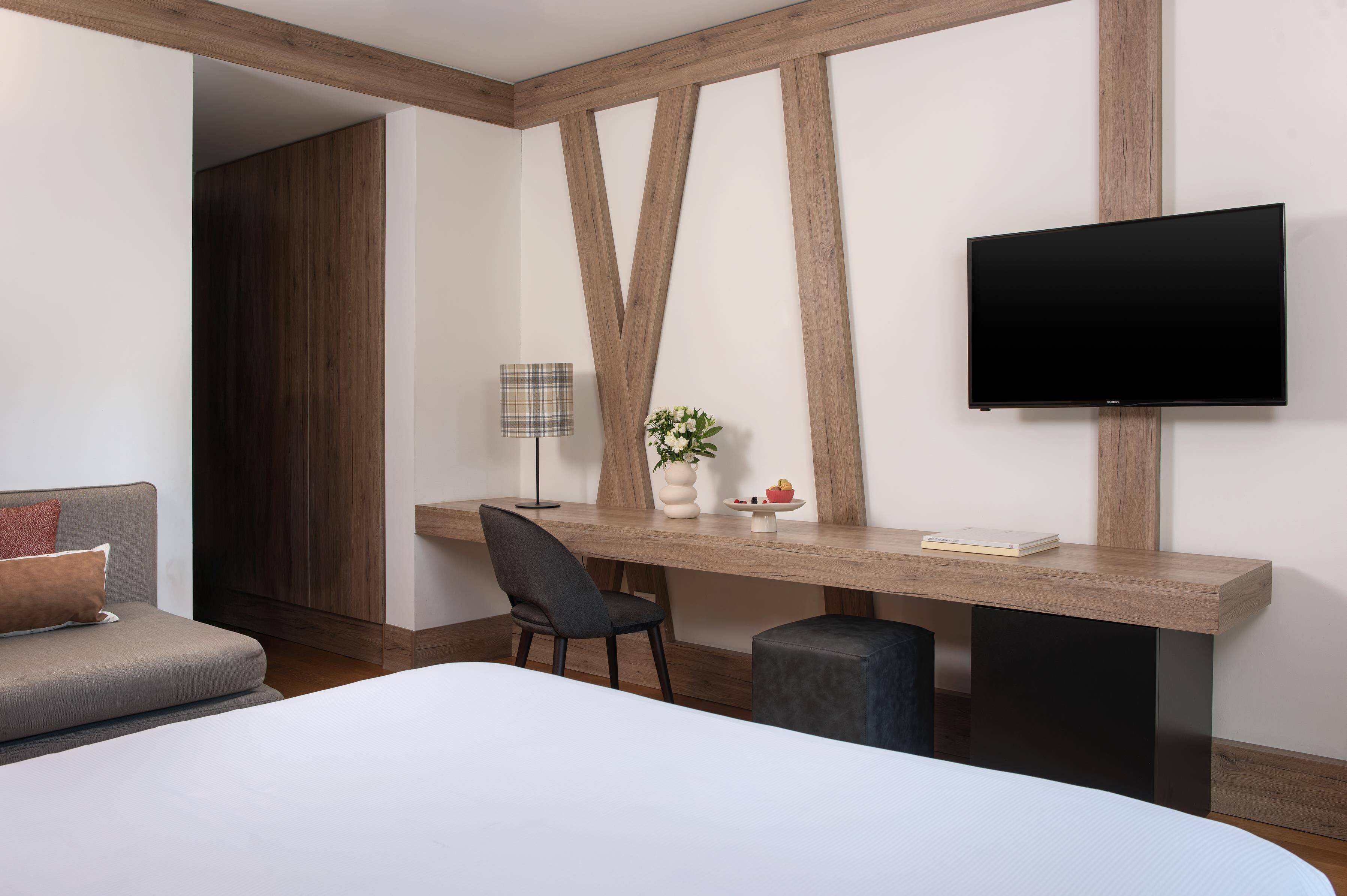 Una camera d'albergo moderna con un letto, una scrivania con una sedia, una TV e un piccolo tavolino con un vaso e un libro.