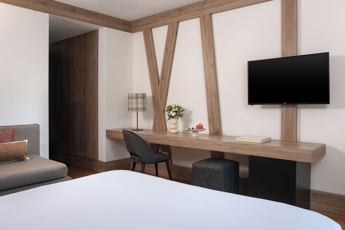 Una habitación de hotel moderna con una cama grande, un escritorio con silla, un televisor de pantalla plana, una lámpara y un jarrón con flores.