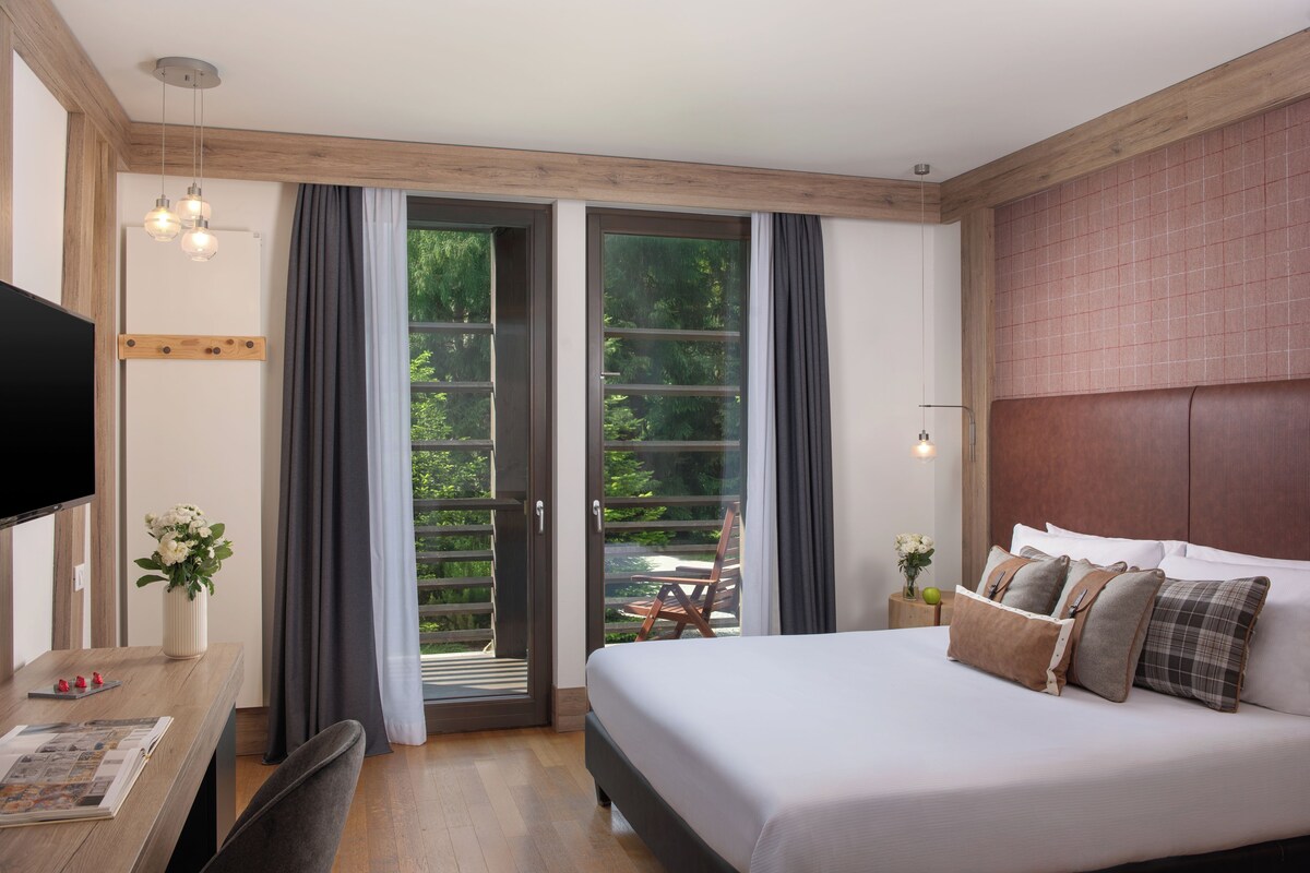 A bedroom with a large bed, a desk with a flat-screen TV, a wooden ceiling, and a sliding glass door leading to an outdoor area.