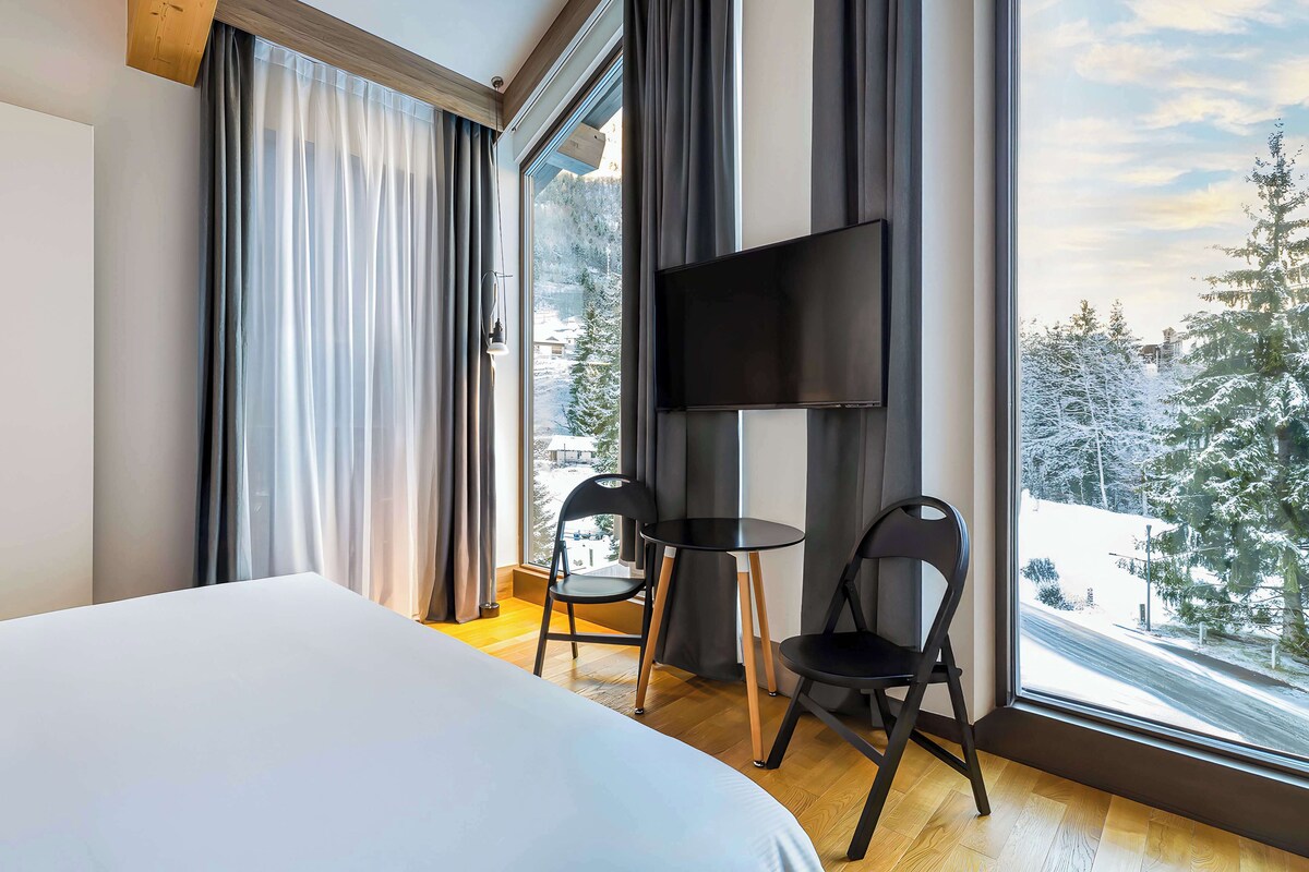 A hotel room with a bed, two chairs, a small table, a TV, and a large window with a view of snow-covered trees.