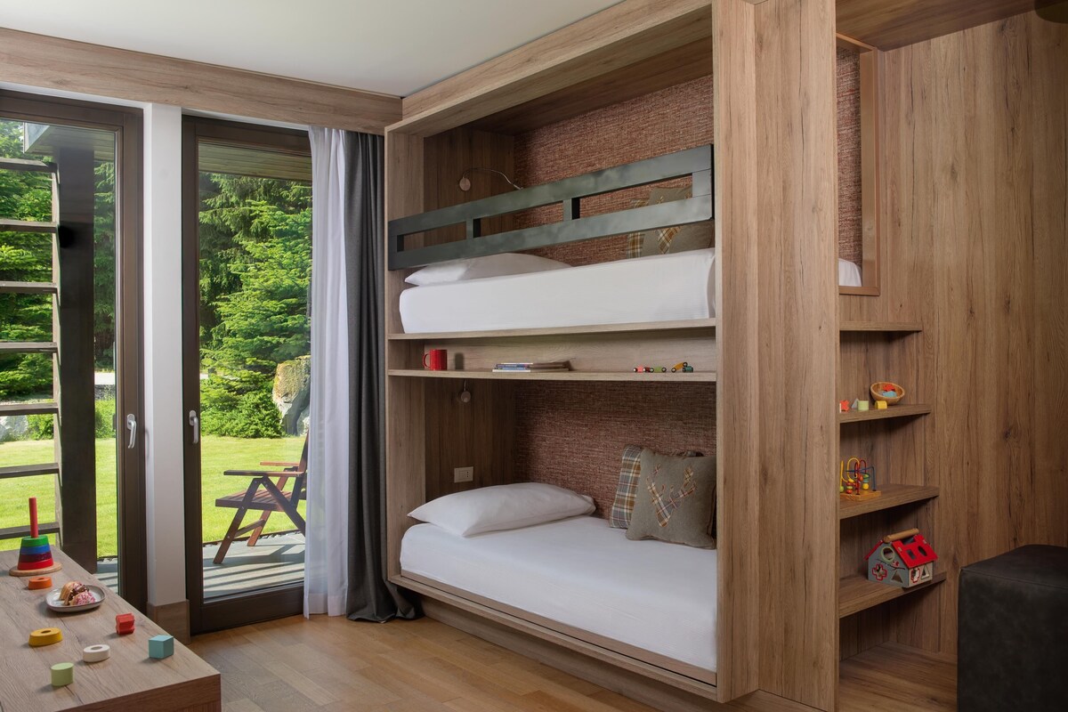 A bunk bed with a wooden headboard, a small table with toys, and a view of a garden through a glass door.