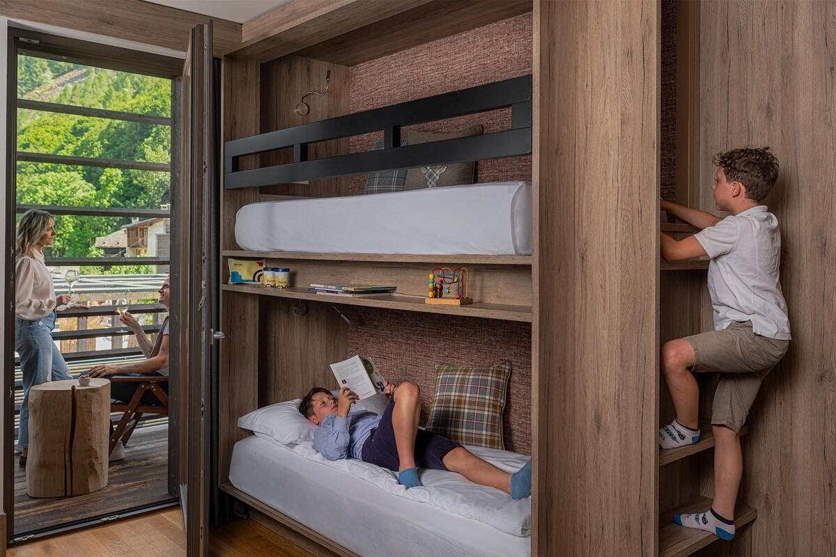 Een stapelbed met een onderbed waar iemand een boek leest, en een bovenbed waar iemand aan het klimmen is. Een derde persoon zit buiten de kamer aan een tafel.