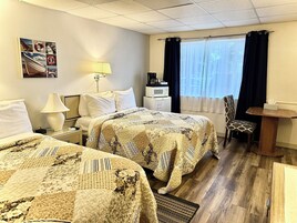 Habitación de hotel con dos camas, un escritorio, una silla, una ventana con cortinas, una lámpara y una obra de arte colgada en la pared.