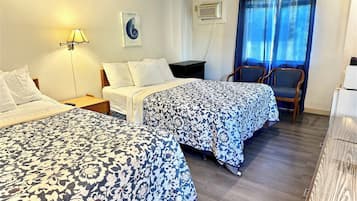Quarto de hotel com duas camas, uma escrivaninha, uma cadeira, um abajur e uma janela com cortinas.