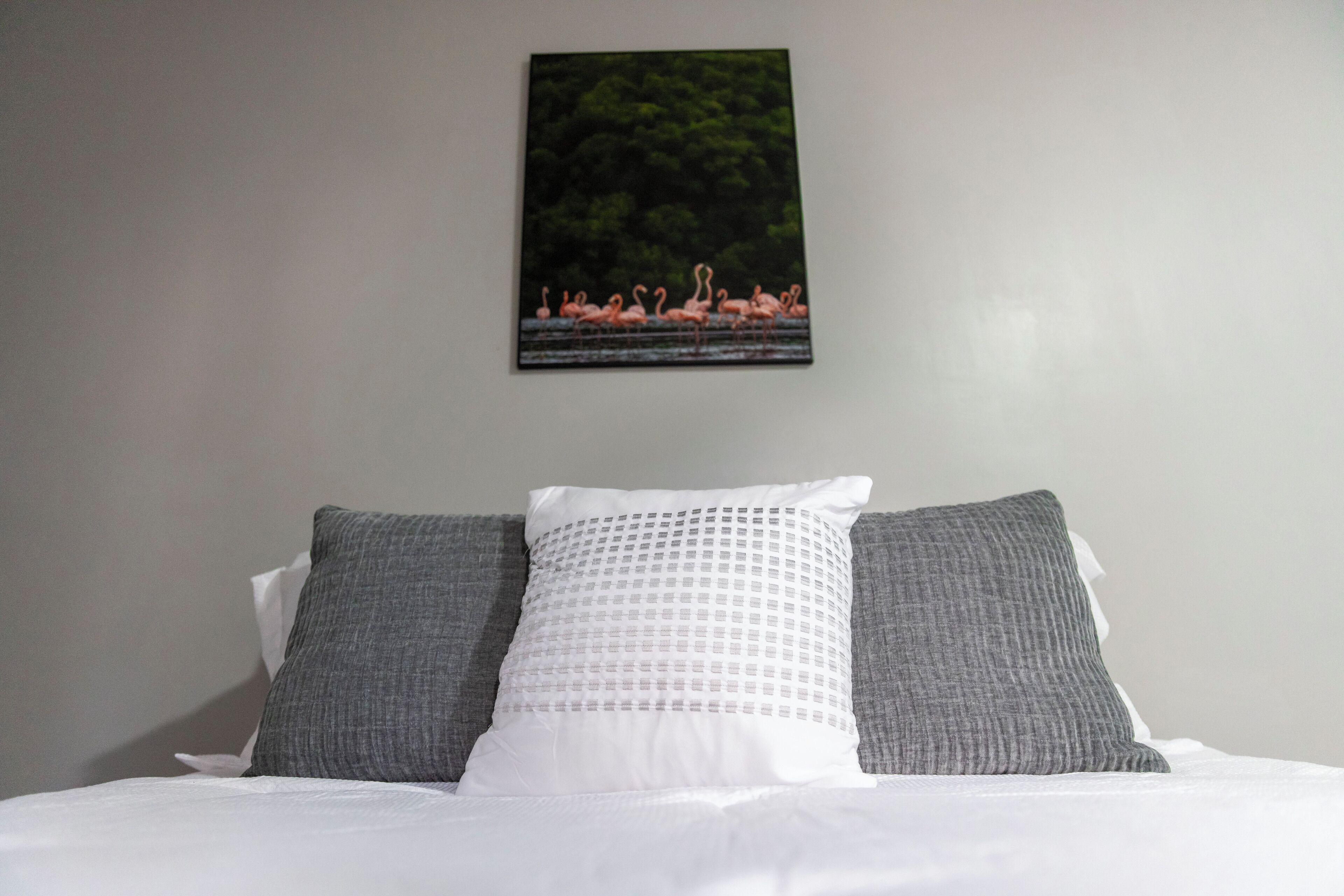 Un lit avec une literie blanche et deux oreillers gris, un tableau représentant des flamants roses accroché au mur, et un mur uni.