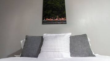 Un lit avec une literie blanche et deux oreillers gris, un tableau représentant des flamants roses accroché au mur, et un mur uni.