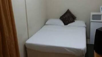 Een kleine eenpersoonskamer met een wit matras, een bruin kussen, een wit nachtkastje en een kleine, witte airconditioningunit aan de muur.