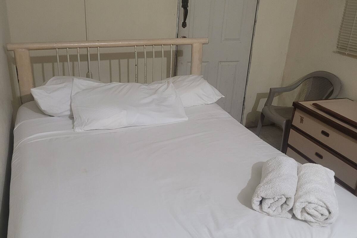 Ein Bett mit weißen Leinentüchern und zwei aufgerollten Handtüchern, ein Holz-Kopfbrett, eine weiße Tür mit einem Schild ‚Nichtraucher‘, ein Plastikstuhl und eine kleine Holzkommode.