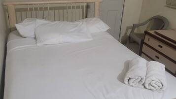 A bed with white linens and two rolled towels, a wooden headboard, a white door with a 'Thank You for Not Smoking' sign, a plastic chair, and a small wooden dresser.