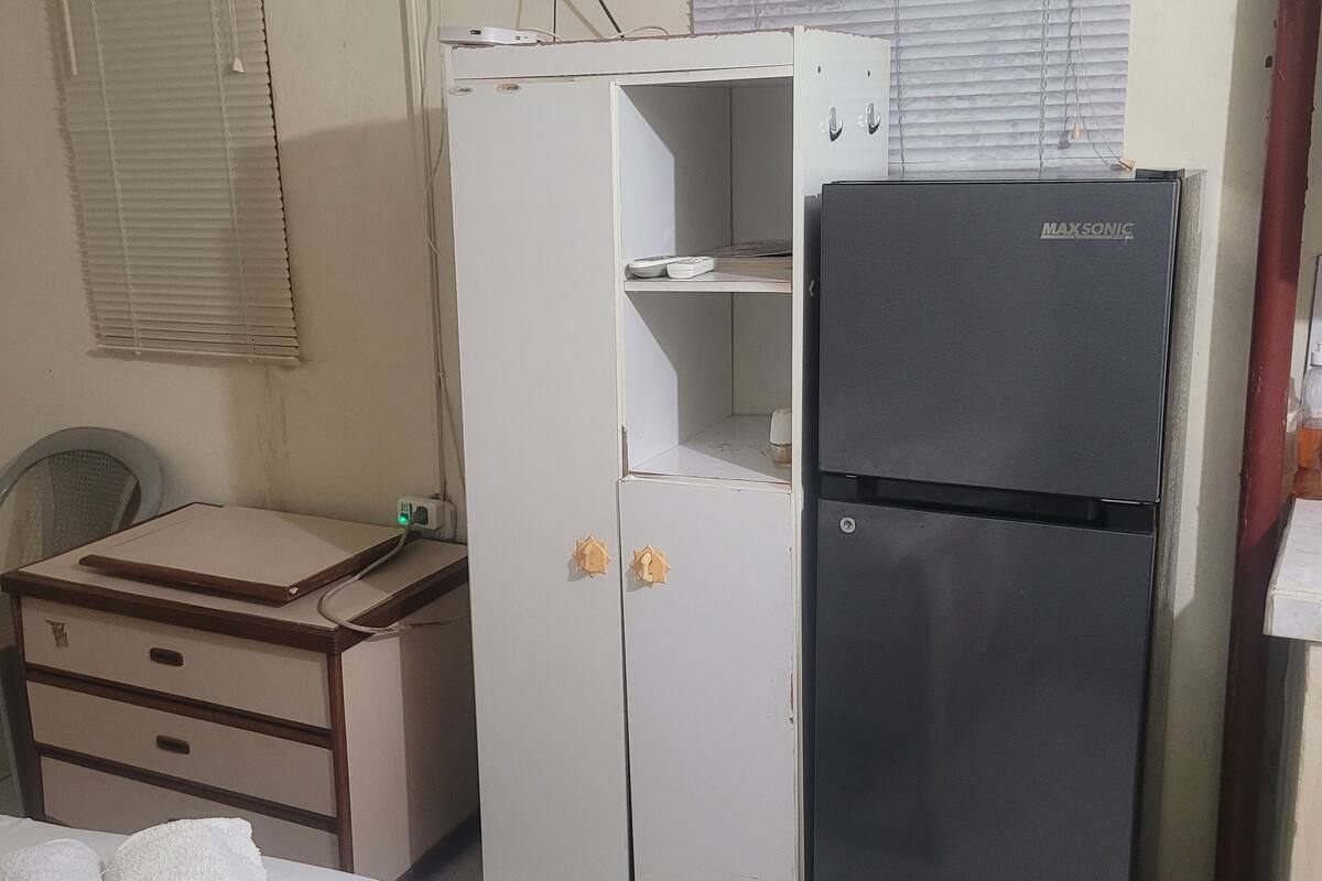 Ein kleiner, kompakter Raum mit einem Bett, einem Schreibtisch, einem Kühlschrank und einem an der Wand befestigten Fernseher.