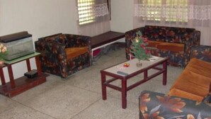 A living room with a sofa, armchair, and a coffee table. There is a television on a stand and a small table with a plant. - Tony's Guest House (Diego Martin)